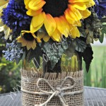 Rustic-style centerpiece featuring sunflowers, blue delphinium, blue pom poms, and gerbera daisies in a yellow-tinted mason jar tied with burlap and twine
