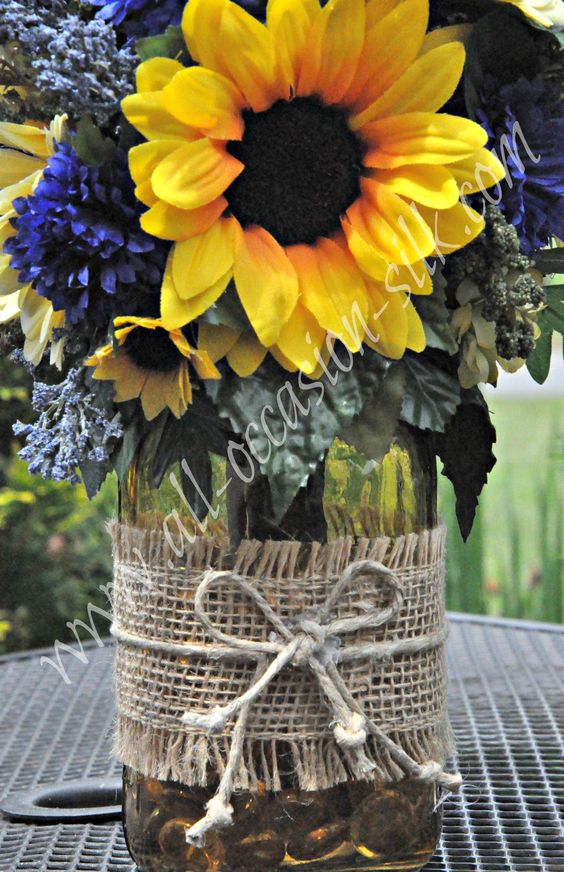 Rustic-style centerpiece featuring sunflowers, blue delphinium, blue pom poms, and gerbera daisies in a yellow-tinted mason jar tied with burlap and twine